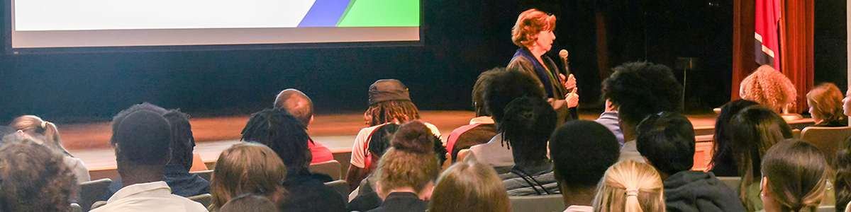 Dr. Janet F. Smith addressing an audience of students in a theater, with a large screen displaying a presentation in the background. The audience consists of diverse individuals seated, some facing the speaker and others turned slightly.
