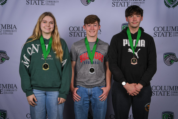 Photo shows a group of three people smiling at the camera with a white background featuring a repeated Columbia State logo pattern.