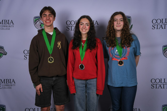 Photo shows a group of three people smiling at the camera with a white background featuring a repeated Columbia State logo pattern.