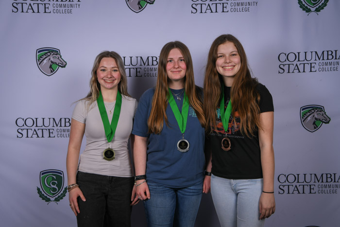 Photo shows a group of three people smiling at the camera with a white background featuring a repeated Columbia State logo pattern.