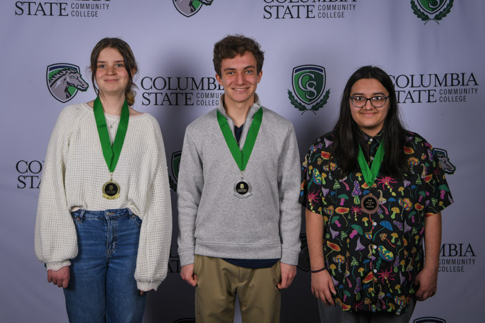 Photo shows a group of three people smiling at the camera with a white background featuring a repeated Columbia State logo pattern.
