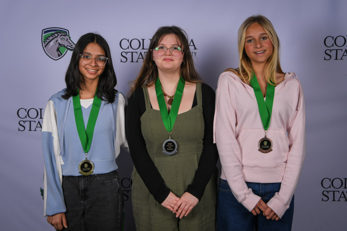 Photo shows a group of three people smiling at the camera with a white background featuring a repeated Columbia State logo pattern.
