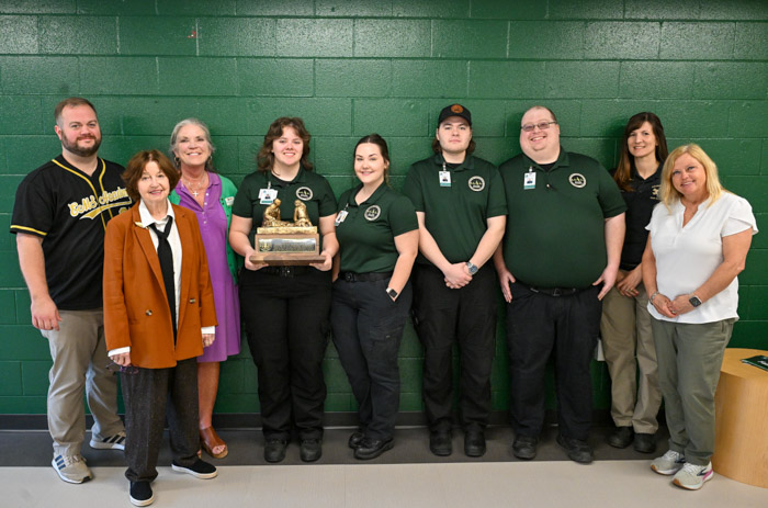 A group of people stand in front of a green wall while holding a wood and metal trophy.