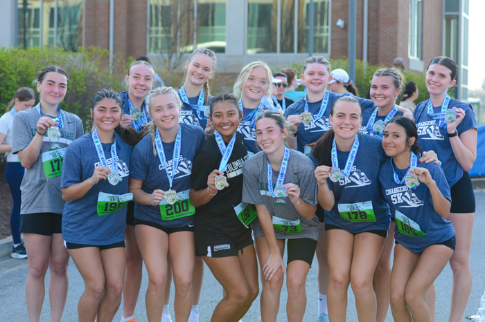 A group of young women hold up medals as they smile got a photo in front of shrubbery.