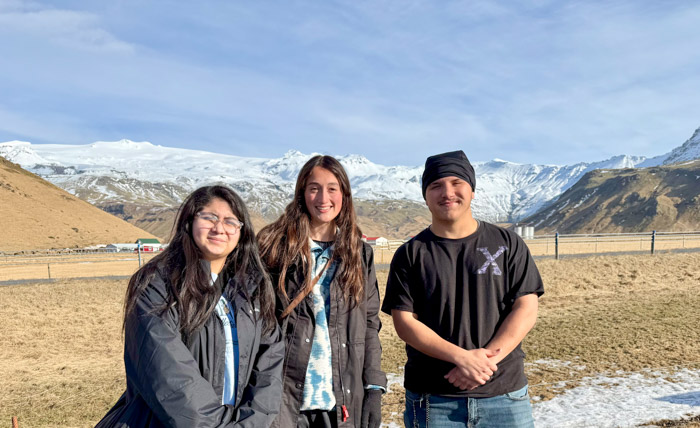 Three people pose for a photo in front of an open landscape with a snow-covered mountain in the background.