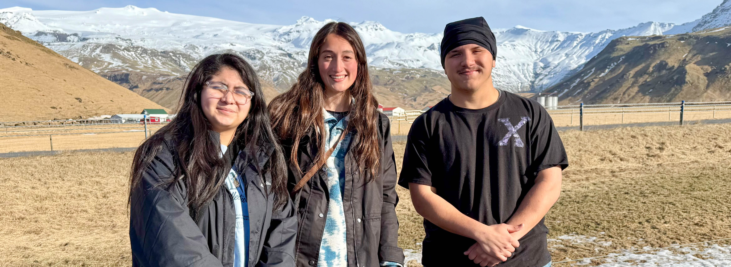 Three people pose for a photo in front of an open landscape with a snow-covered mountain in the background