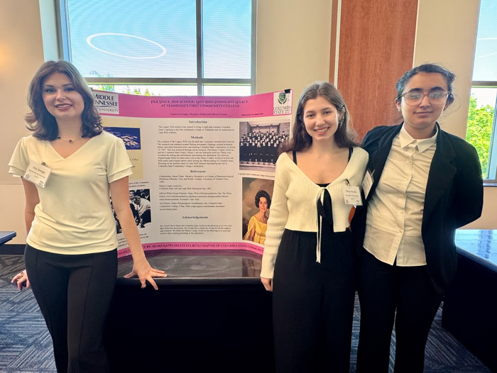 Three women stand in front of a poster display and smile.