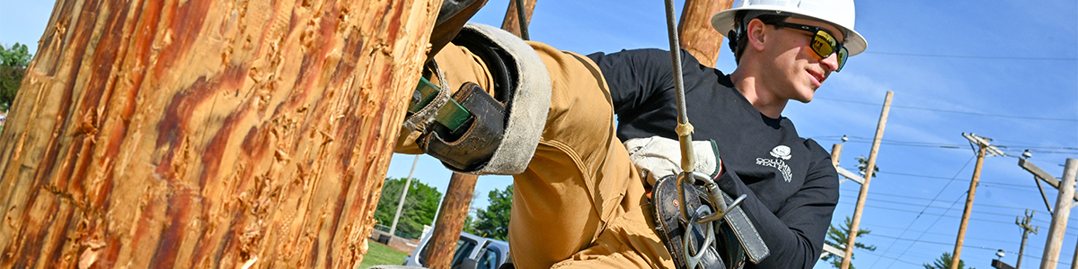 A lineman in a hard hat and safety glasses works on a utility pole, wearing climbing gear against a blue sky with power lines and poles in the background.